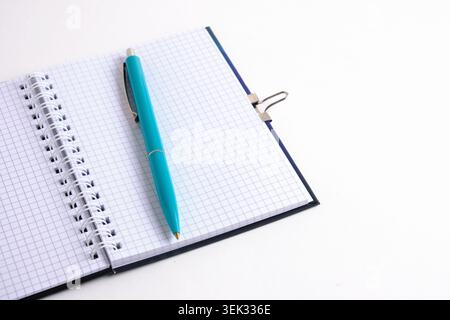 Der türkisfarbene Stift liegt schräg auf einem offenen spiralförmigen quadratischen Notizblock auf weißem Hintergrund. Angewinkelte Ansicht Stockfoto