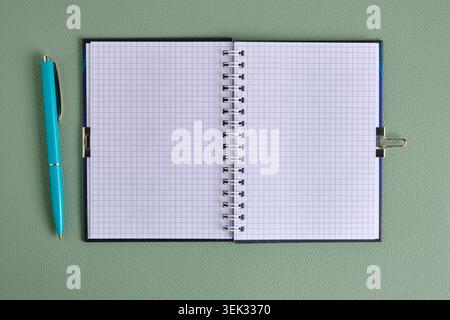 Das offene leere quadratische Notizbuch liegt neben dem blauen Stift auf einer grünen, strukturierten Lederoberfläche. Draufsicht Stockfoto