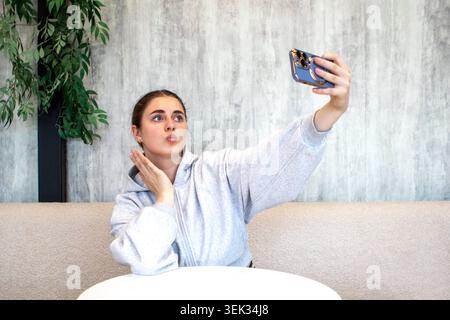 Eine junge Frau, die ein Selfie in einem Café macht, sich die Lippen kneift und mit einem Smartphone posiert Stockfoto