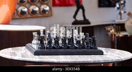 Schach mit schwarzen und silbernen Stücken auf einem runden Marmortisch auf einem Flohmarkt, umgeben von verschiedenen dekorativen Gegenständen und Skulpturen Stockfoto