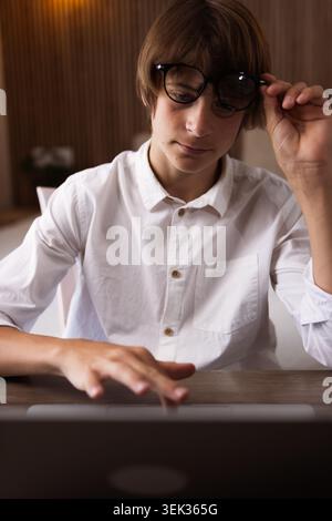 Junger Mann in weißem Hemd und Brille, der sich auf einen Laptop konzentriert, von zu Hause aus studiert und arbeitet, für Online-Lernen, Fernarbeit und digitales Licht Stockfoto