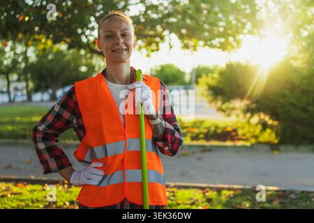 Weibliche Sanitärangestellte in einer gut sichtbaren Weste und Handschuhen, die in einem Park stehen und einen Besen halten, um öffentliche Räume sauber zu halten und zu fördern Stockfoto
