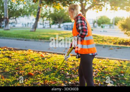 Freiwillige Frau trägt eine orangefarbene Sicherheitsweste und weiße Handschuhe, die aktiv Herbstlaub in einem grünen öffentlichen Park harkt und so zum Gemeindedienst beiträgt Stockfoto
