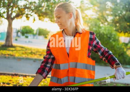 Frau in orangefarbener Sicherheitsweste und Handschuhen sammelt Müll in einem sonnigen öffentlichen Park und meldet sich freiwillig für die Gemeindereinigung, um die Gegend zu verschönern und zu schützen Stockfoto