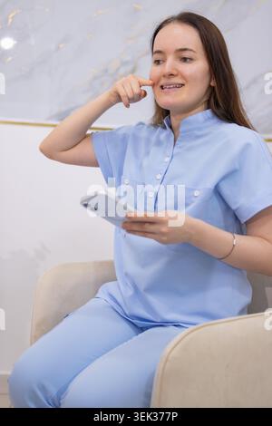 Junge Frau in medizinischer Uniform sitzt bequem, hält ein Smartphone, berührt ihr Gesicht, während sie wegblickt, repräsentiert digitale Kommunikation A Stockfoto