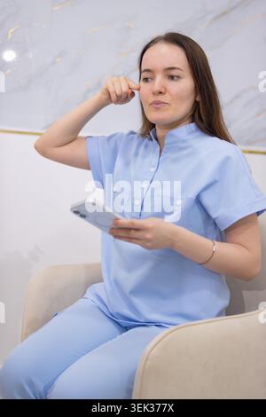 Weibliche Gesundheitsberaterin in blauen medizinischen Peelings, die in einer modernen Klinik sitzen und ihr Gesicht in der Nähe ihres Tempels berühren, während sie ihr Smartphone, r Stockfoto