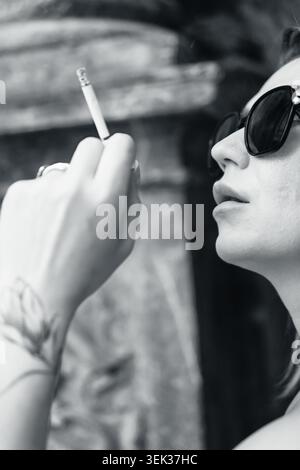 Stilvolle Frau mit dunkler Sonnenbrille und einer brennenden Zigarette mit einem Arm-Tattoo, das in einem auffälligen Schwarz-weiß-Porträt festgehalten wird, das an einen erinnert Stockfoto