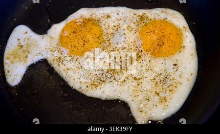 Blick aus einem hohen Winkel auf zwei sonnige Spiegeleier, gewürzt mit getrockneten grünen Kräutern und Gewürzen, die in einer schwarzen Pfanne gekocht werden. Die glänzenden gelben Eigelbe sind es Stockfoto