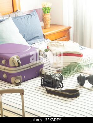 Helles Schlafzimmer mit violettem Koffer, gefalteter Kleidung, Kamera, Kopfhörern und Palmblatt für die Vorbereitung des Sommerurlaubs. Stockfoto