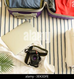Blick von oben auf einen offenen lila Koffer mit Kleidung, Laptop, Kamera und Palmblatt auf einem gestreiften Bett für den Sommerurlaub. Stockfoto