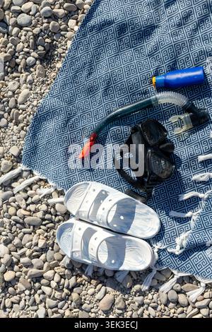 Strandurlaubs-Essentials auf einem Handtuch über Kieselstrand, einschließlich Schnorchelmaske, Tube, Sonnencreme und Sommer-Accessoires während der sonnigen Küste Stockfoto