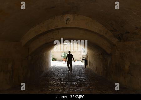 Touristen, die unter einem alten Bogengang in einer mittelalterlichen Stadt vorbeifahren, dramatische Hintergrundbeleuchtung und Kontrast schaffen ein Reise- und Erkundungskonzept Stockfoto