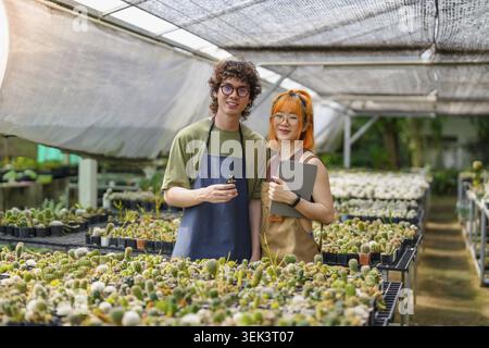 Glückliche junge asiatische Landbesitzer und Landbesitzer, die Kaktuspflanzen in grünen Gewächshäusern für kleine Unternehmen anbauen Stockfoto