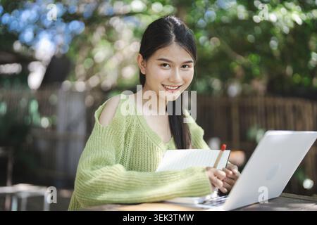 Schöne junge asiatische Studenten studieren im Freien im Öko-Café, schreiben auf einem Notebook und arbeiten an einem Laptop. Akademische Forschung mit Outdoor-Arbeiten Stockfoto