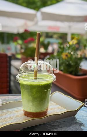 Matcha Smoothie in Plastikbecher mit Strohständern auf Kaffeetablett neben Gartenpflanzen und Sonnenschirmen. Konzept von Matcha-Drink, Wellness, Café-Erfrischung Stockfoto