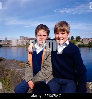 Zwei Jungen posieren in Cork, Irland. Im Hintergrund ist eine historische Stadtansicht mit einer Burg und einem Fluss zu sehen. 1985. Stockfoto
