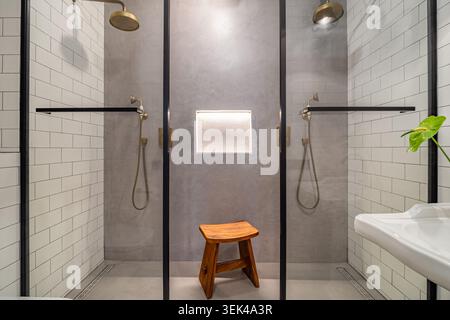Modernes Badezimmer mit zwei Duschkabinen aus Glas, einem Holzhocker und einer kleinen grünen Pflanze. Die Wände sind in weiß- und Grautönen gefliest. Stockfoto