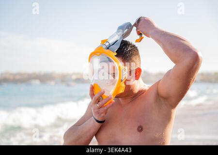 Der kaukasische Mann passt die Vollgesichts-Schnorchelmaske an einem Strand mit Meereswellen und nasser Haut während des sonnigen Sommerurlaubs an. Schnorcheln und Erholung im Meer. Stockfoto