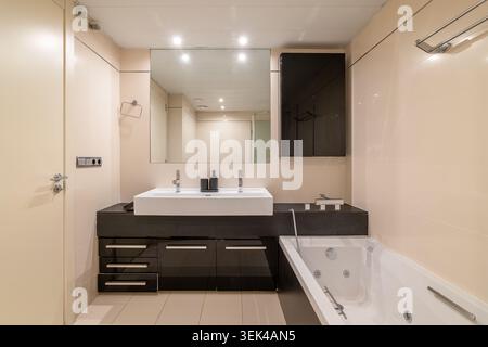 Modernes Bad mit elegantem Design. Verfügt über ein großes weißes Waschbecken, schwarze Schränke, Badewanne und helle Beleuchtung. Klare und minimalistische Ästhetik. Stockfoto