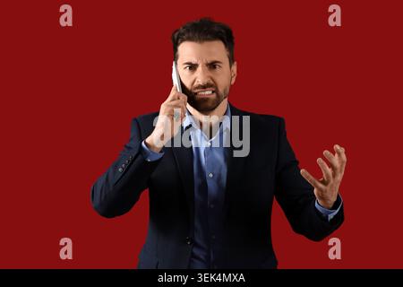 Wütender Geschäftsmann, der mit dem Handy auf rotem Hintergrund spricht Stockfoto