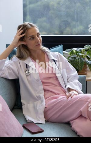 Weibliche Krankenschwester in rosafarbenen Schutten, die auf der Couch sitzen, sich beugen, zum Fenster, zum Stethoskop und zum Telefon blicken Stockfoto