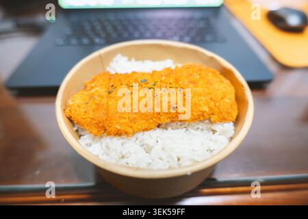 Knusprige panierte Reisschale mit Schnitzel auf Holzschreibtisch mit Laptop im Hintergrund Stockfoto