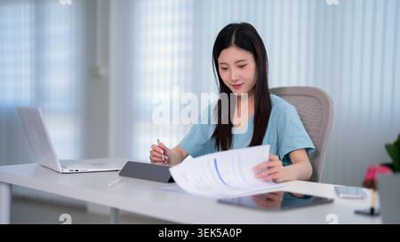 Asiatische Frau in blau schrubbt Dokumente am Schreibtisch mit Laptop, die von Heimbüro-Gesundheitsfachkraft arbeitet Stockfoto
