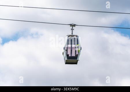 Eine moderne Seilbahnkabine hängt an zwei Stahllinien vor einem hellen, bewölkten Himmel. Die isolierte Gondel schafft ein klares Bild von Fahrt und Transport. Stockfoto