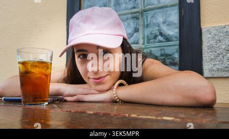 Junge Frau mit rosa Kappe, die sich neben einem Glas Eistee an einem Café-Tisch im Freien entspannt. Stockfoto