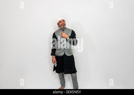 Indischer Mann in traditioneller Kleidung, darunter eine graue und schwarze Weste, eine dunkle Hose und eine weiße Mütze. Er hält ein Smartphone und posiert aga Stockfoto