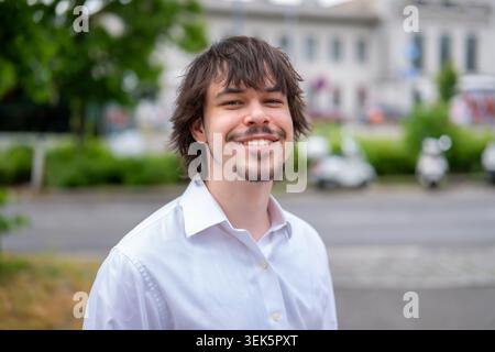 Junger lächelnder Mann in weißem Hemd, der im Sommer in Österreich auf der Straße posiert Stockfoto