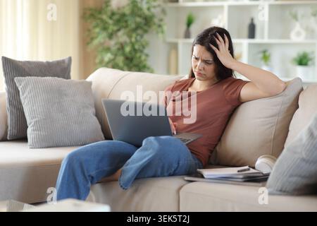 Besorgter junger Student, der schlechte Nachrichten auf einem Laptop liest, der zu Hause auf einem Sofa sitzt Stockfoto