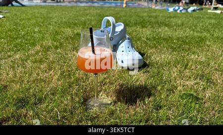 Foto von hellblauen Strandschuhen und einem orangefarbenen Cocktail auf hellgrünem Gras mit sichtbarem Swimmingpool im Hintergrund. Weitwinkel-Aufnahmen Stockfoto