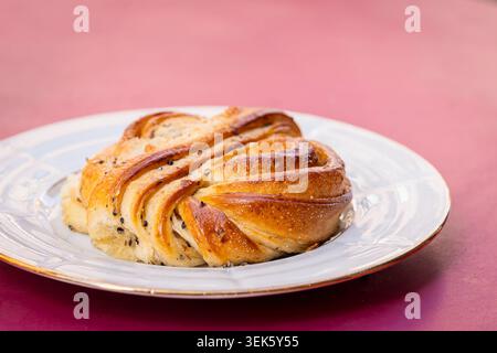 Frisch gebackenes skandinavisches Kardamombrötchen auf einem weißen Teller mit goldenen geflochtenen Schichten und aromatischer Gewürzfüllung vor einem weichen rosa Hintergrund. Stockfoto