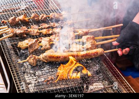 Aus nächster Nähe greifen Sie mit der Hand nach schrägen Würstchen neben Schweinefleisch auf Spießen, die auf einem Metallgrill mit Flammen an einem Lebensmittelstand im Ueno Park kochen. Stockfoto