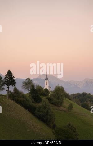 Kleiner Kirchturm auf einem grünen Hügel mit Bäumen, Bergen und einem weichen, pastellfarbenen Himmel in der Abenddämmerung. Stockfoto