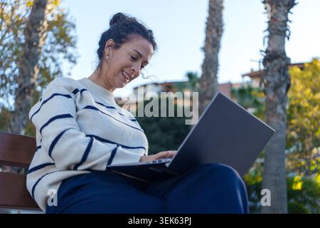 Frau, die auf einer Bank sitzt und glücklich auf ihrem Laptop schreibt. Sie arbeitet ferngesteuert und bleibt mit Technologie im Freien in Verbindung Stockfoto