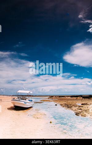 Ein kleines weißes Boot ruht am Sandstrand des Ujung Genteng Beach, Indonesien. Das Boot ist festgebunden, mit einem Baldachin, das Schatten spendet. Um sie herum Stockfoto