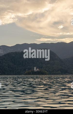 Ruhiges Seewasser mit Wellen, bewaldete Berge, eine kleine Kirche am Ufer und ein weicher bewölkter Himmel bei Sonnenuntergang. Stockfoto