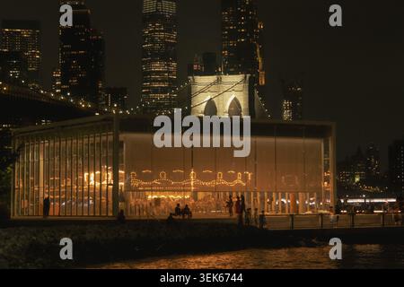 Nachtansicht auf einen beleuchteten Glaspavillon am Wasser mit städtischen Wolkenkratzern und einer Brücke im Hintergrund, die das urbane Nachtleben und die Reflexionen zeigt Stockfoto