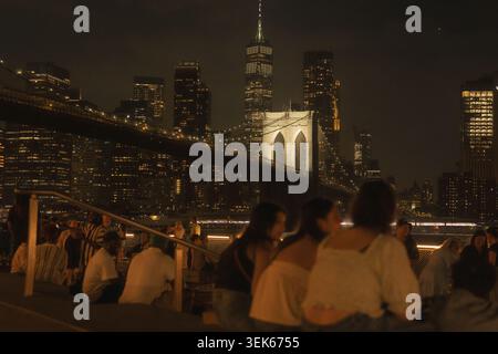 Die Leute sitzen und unterhalten sich nachts in der Nähe einer beleuchteten Brücke mit einer Skyline der Stadt im Hintergrund Stockfoto