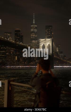 Verschwommene Person, die nachts die brooklyn Bridge und die Skyline von manhattan vom Ufer aus fotografiert. Stockfoto