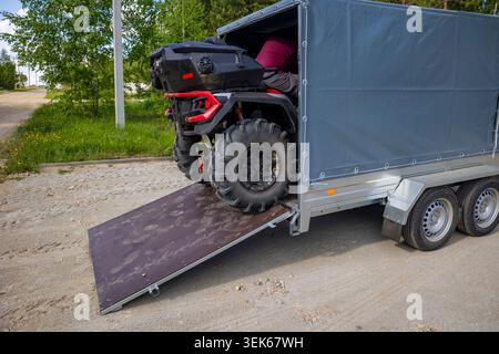 Fahren eines schwarzen Quads in einen grauen, überdachten Lastenanhänger auf einer offenen Sperrholzrampe. Konzept: Transportlogistik Stockfoto