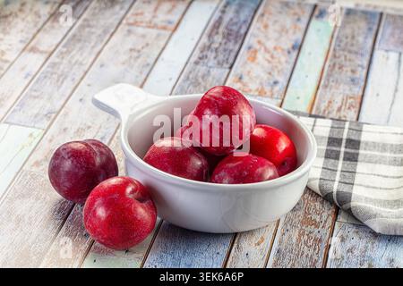 Ein Stillleben aus leuchtenden roten Pflaumen in einer weißen Schüssel. Auf einer rustikalen, mehrfarbigen Holzoberfläche mit einer karierten Serviette. Natürliches Licht Highlights f Stockfoto