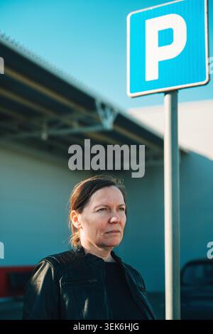 Eine ernsthafte Frau mittleren Alters steht auf einem städtischen Parkplatz unter einem blauen Parkschild, trägt eine Lederjacke und sieht nachdenklich aus. Natürliches Dayl Stockfoto
