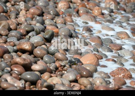 Ocean Wave Foam, der über nassen bunten Kieseln am Rocky Beach gespült wird Stockfoto