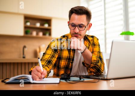 Ein fokussierter junger Mann macht Notizen in einem Notebook, während er an seinem Laptop arbeitet, organisiert Informationen sorgfältig und plant seine nächsten Schritte. Stockfoto