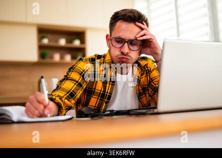 Ein Mann in einem gelben karierten Hemd schreibt in einem Notizbuch, während er sich Inhalte auf seinem Computerbildschirm ansieht. Stockfoto