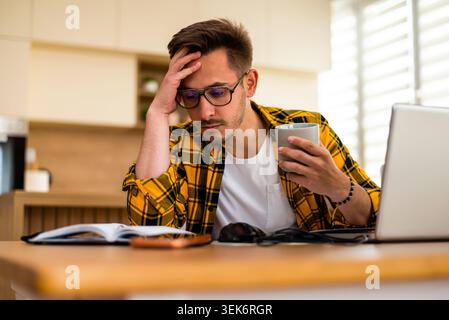 Der junge Mann arbeitet von zu Hause aus, hält eine Tasse Kaffee in der Hand und schaut auf den Notizblock. Stockfoto