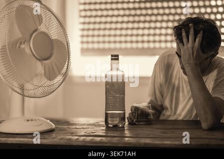 Ein Mann, der unter extremer Hitzeermüdung und Unwohlsein leidet und sich im heißen Sommer mit Trinkwasser versorgt, mit einem elektrischen Ventilator, der ihn abkühlt. Stockfoto